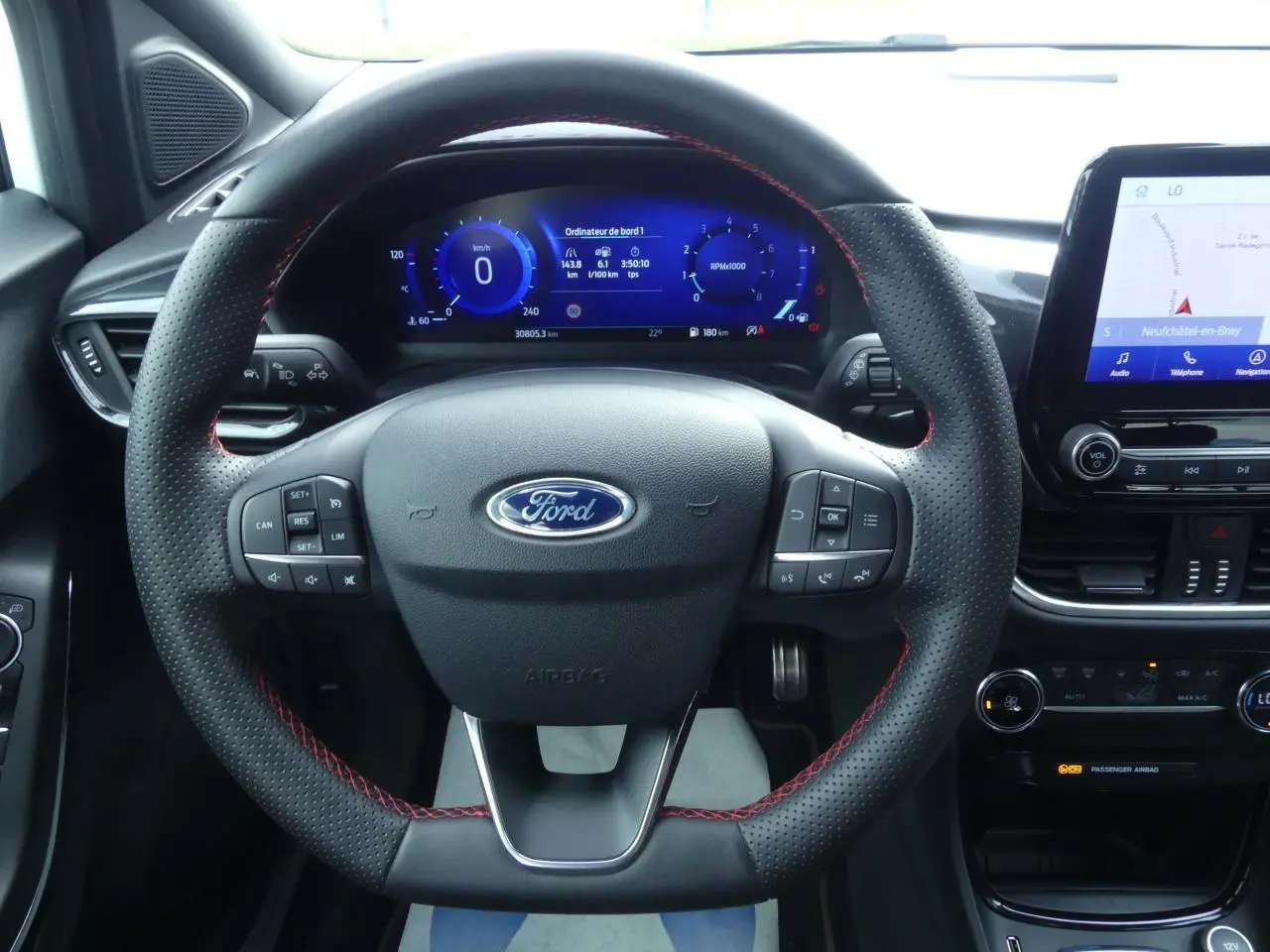 Vue intérieure frontale du volant noir perforé avec surpiqûres rouges du Ford Puma blanc, tableau de bord numérique et écran tactile central visibles.