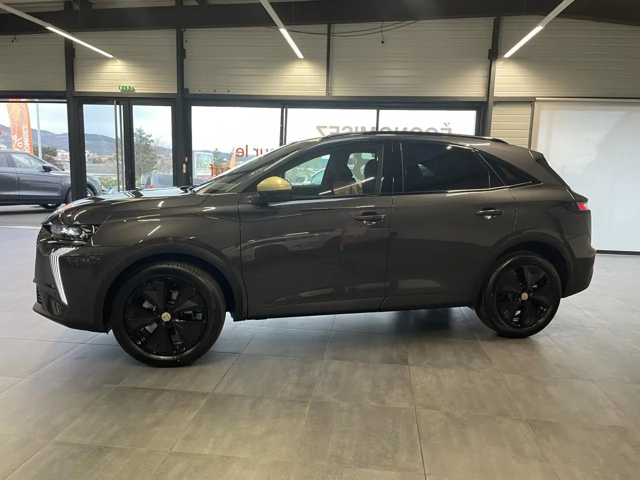 DS7 Crossback gris titanium en profil droit, jantes noires et rétroviseurs dorés satinés visibles en intérieur showroom.