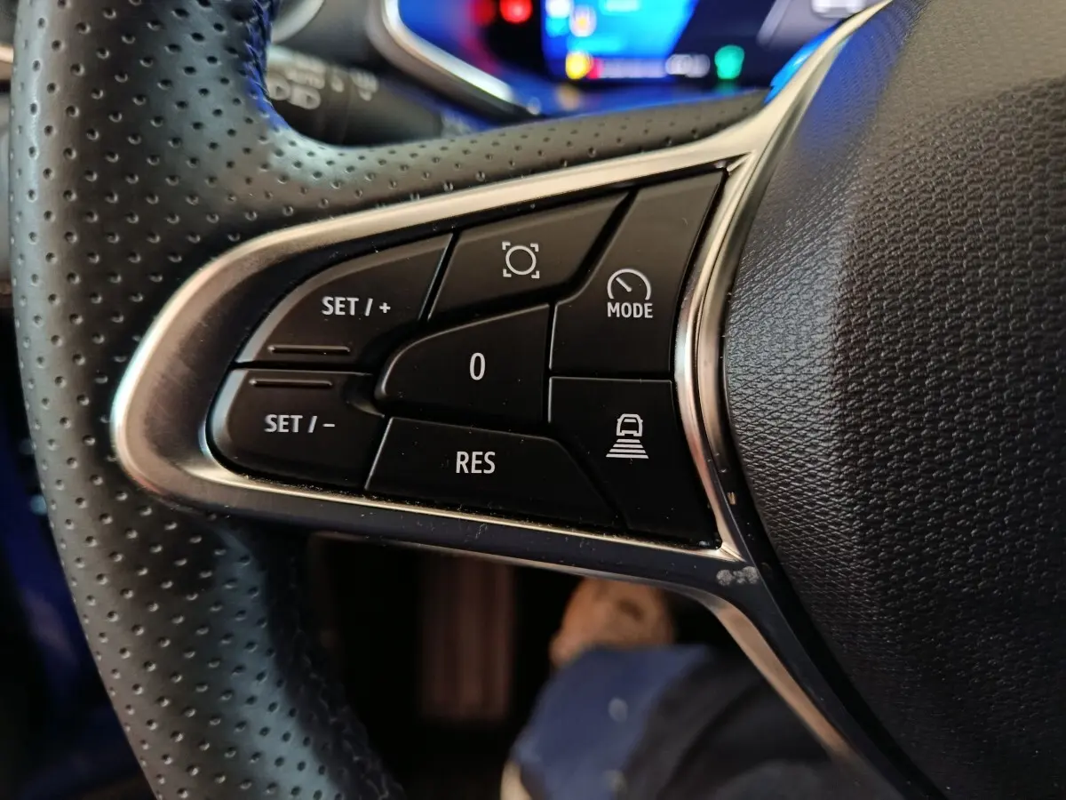 Gros plan sur les commandes noires du volant perforé du Renault Symbioz 2025, version hybride bleu visible en arrière-plan.