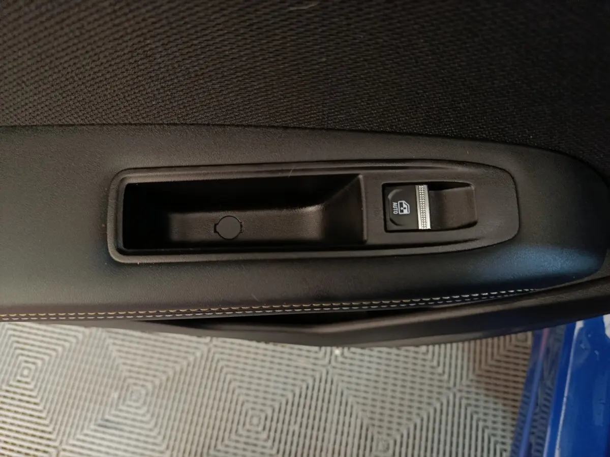 Gros plan sur la commande de lève-vitre côté conducteur, garniture noire avec surpiqûres blanches, porte bleu visible en bas.