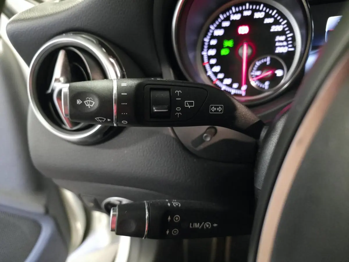 Détail du commodo gauche noir avec commandes d’essuie-glace et régulateur, tableau de bord partiellement visible d’une Mercedes GLA blanche.