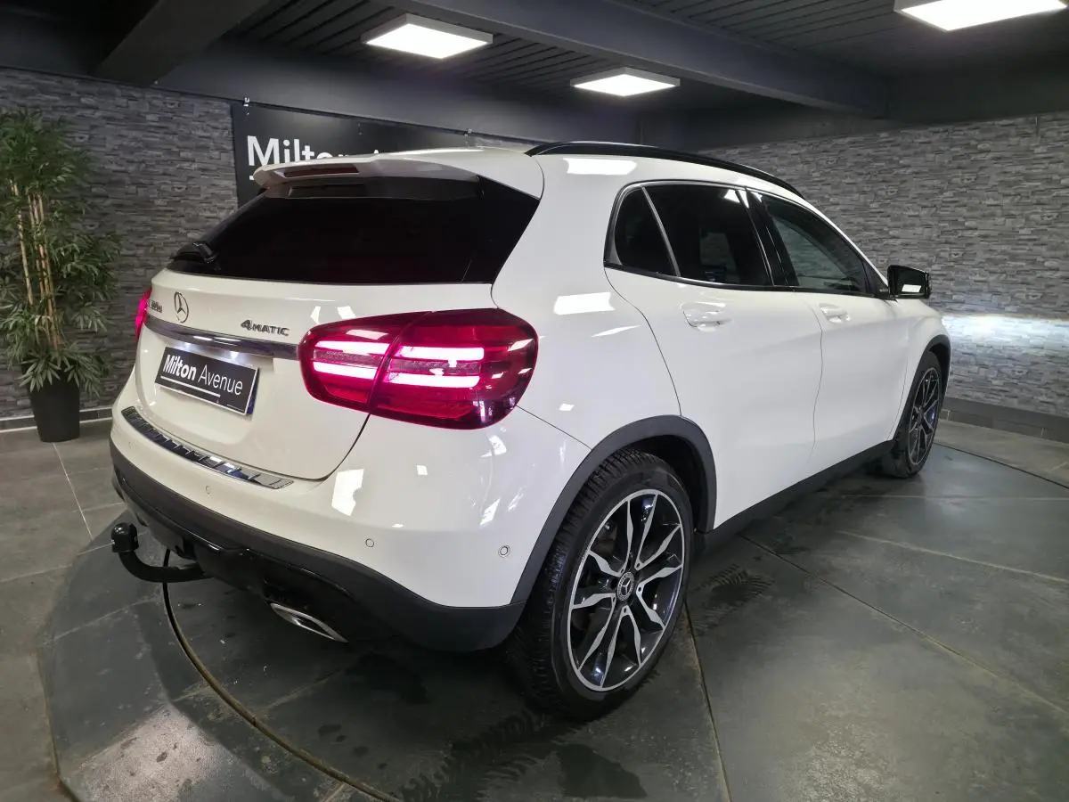 Vue 3/4 arrière droite d’un Mercedes GLA 220 d blanc avec jantes alliage 19’’ et attelage démontable en intérieur showroom.