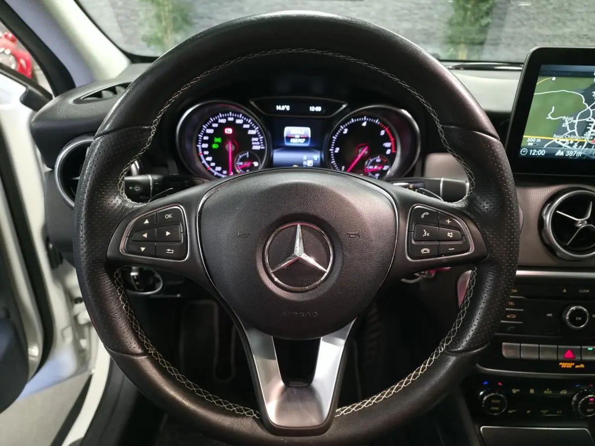 Vue centrée sur le volant en cuir perforé avec surpiqûres contrastées du Mercedes GLA blanc, tableau de bord et écran de navigation visibles.