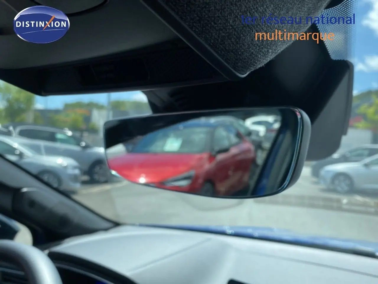 Rétroviseur intérieur montrant un reflet flou d’une voiture rouge, tableau de bord gris et ciel bleu visible.