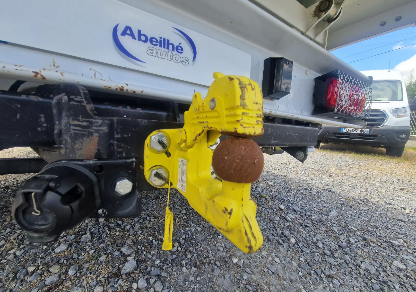 Gros plan sur l'attelage jaune et rouillé à l'arrière droit d'un Citroën Jumper blanc avec benne et ridelles grillagées.
