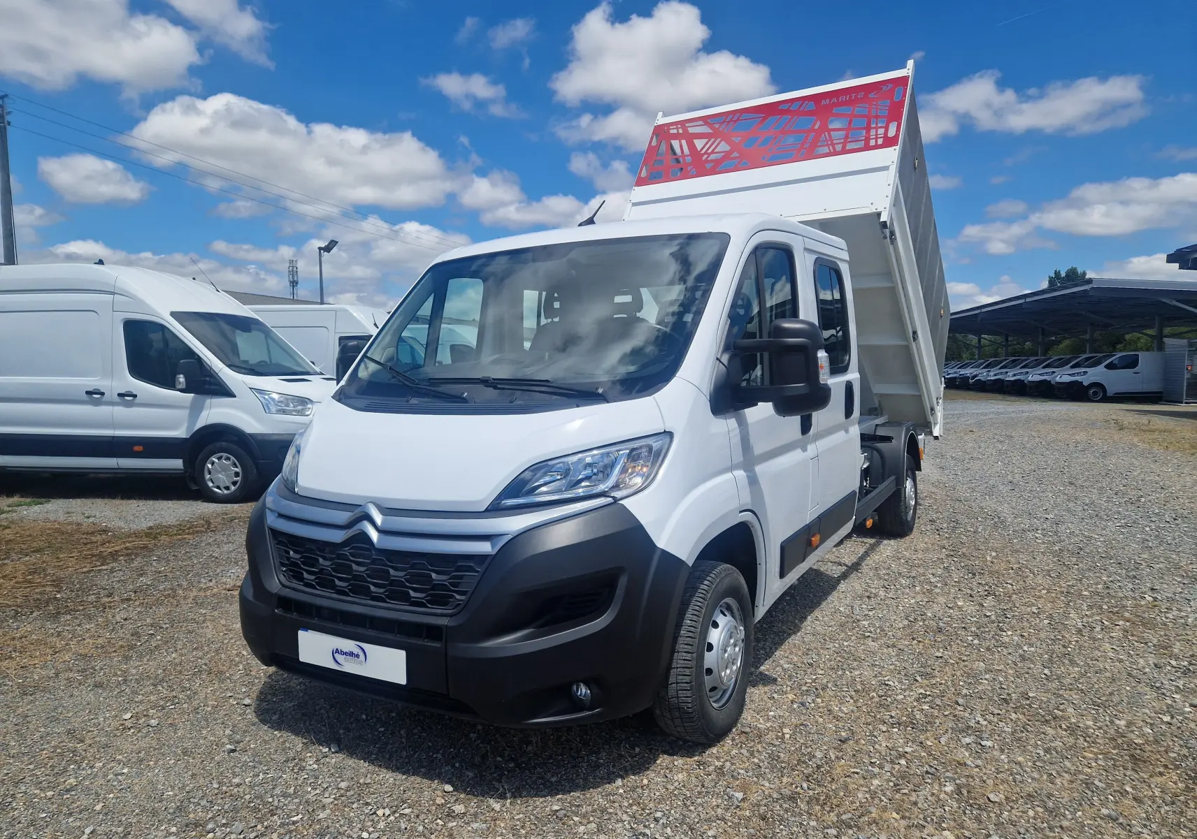 Vue 3/4 avant droite d'un Citroën Jumper blanc avec benne relevée et ridelles grillagées rouges sur un terrain extérieur.