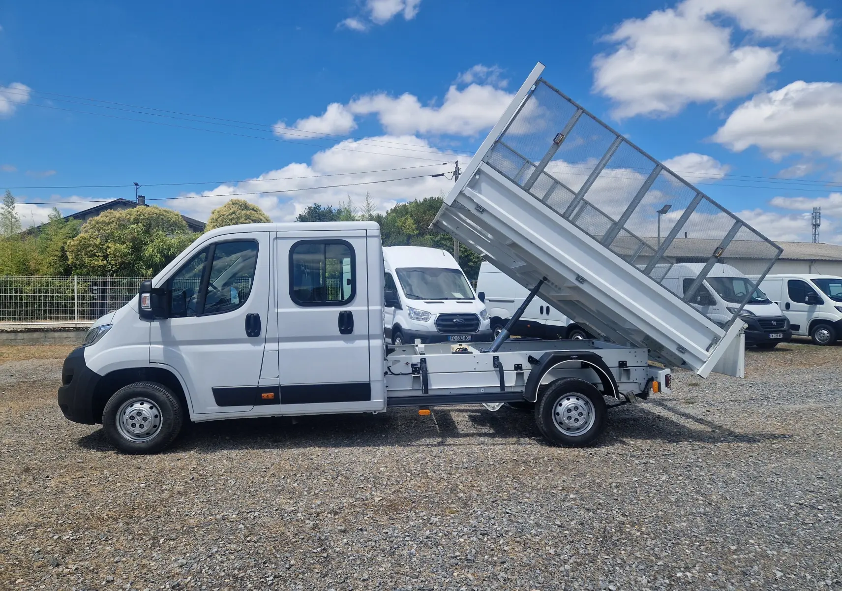 Vue latérale gauche d’un Citroën Jumper blanc avec benne relevée et ridelles grillagées, sur un terrain extérieur.