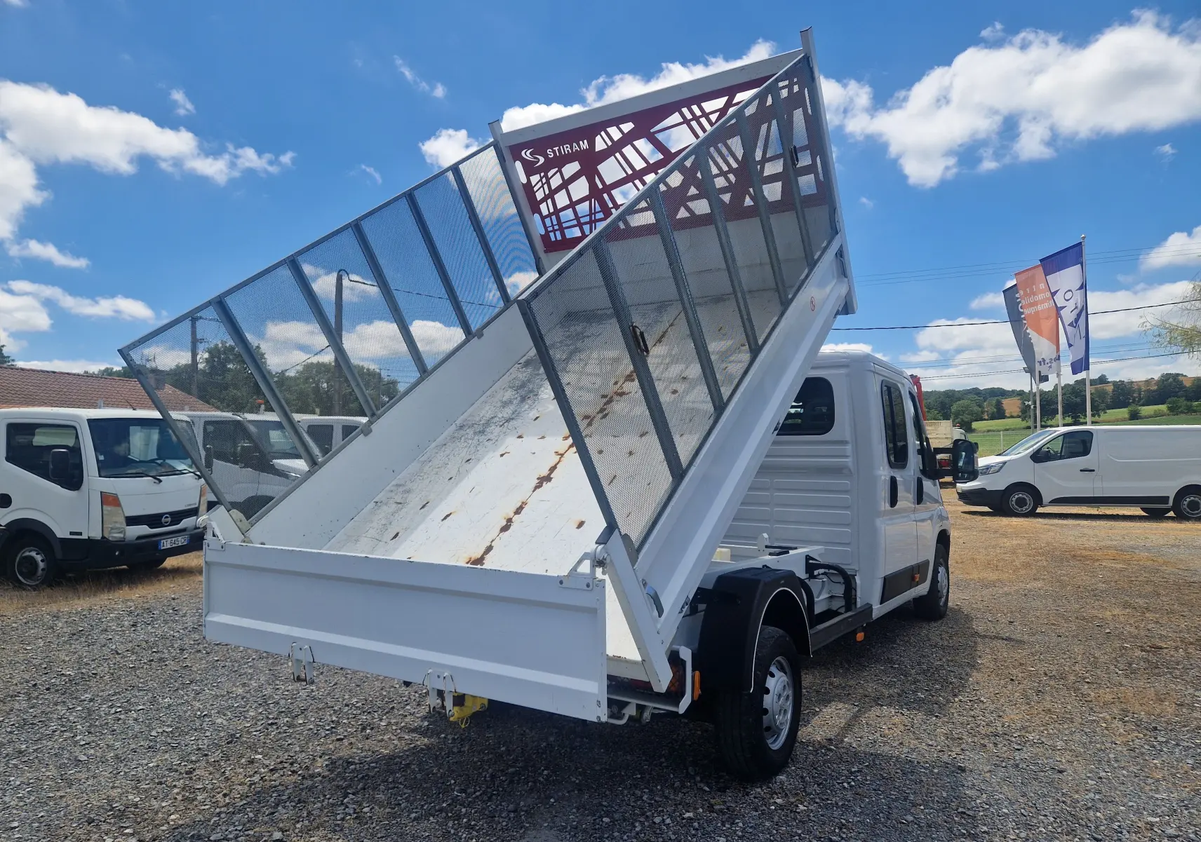 Vue 3/4 arrière droite du Citroën Jumper blanc avec benne relevée et ridelles grillagées sur fond de ciel bleu.