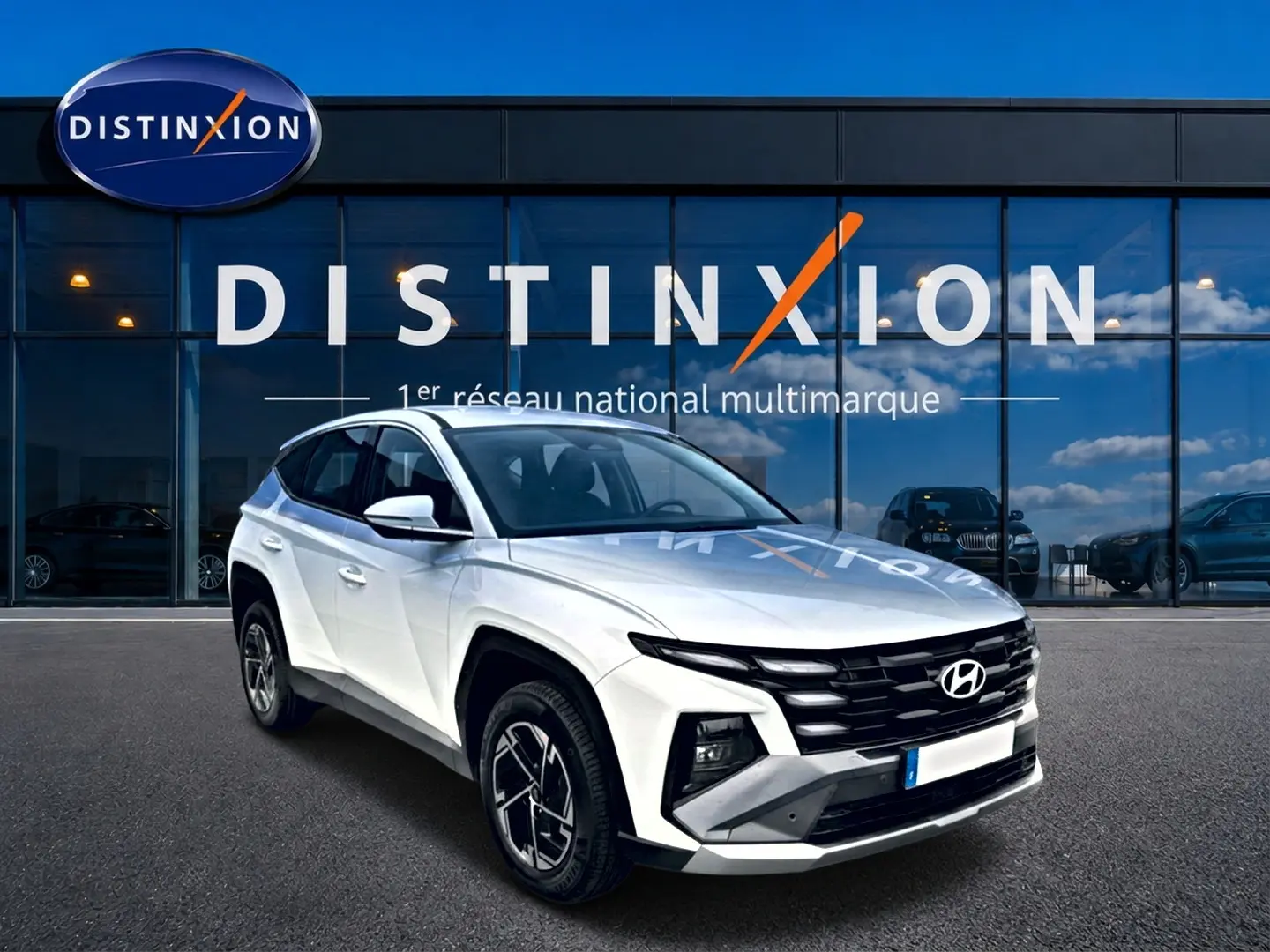 Vue 3/4 avant d'un Hyundai Tucson blanc 2026 avec calandre noire et jantes alliage 17 pouces devant un showroom Distinxion.