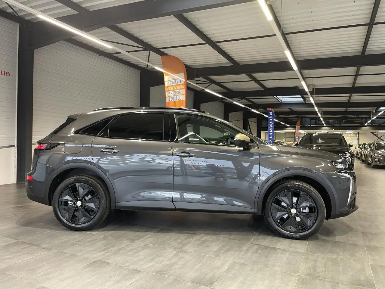 Vue latérale droite du DS7 Crossback gris titanium avec jantes noires et barres de toit noir brillant en intérieur showroom.