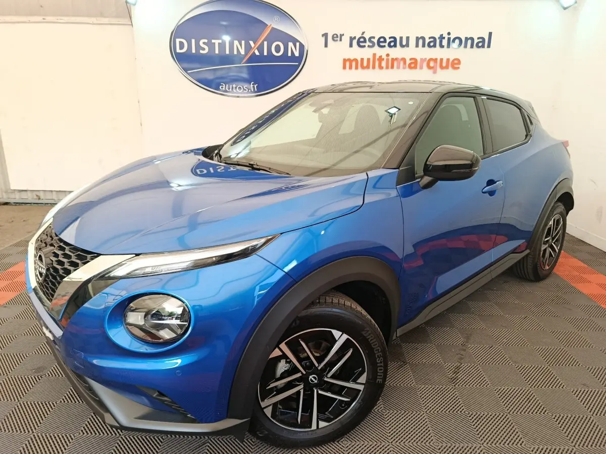 Nissan Juke bleu en 3/4 avant droit, avec toit noir et jantes alliage bicolores visibles en intérieur showroom.
