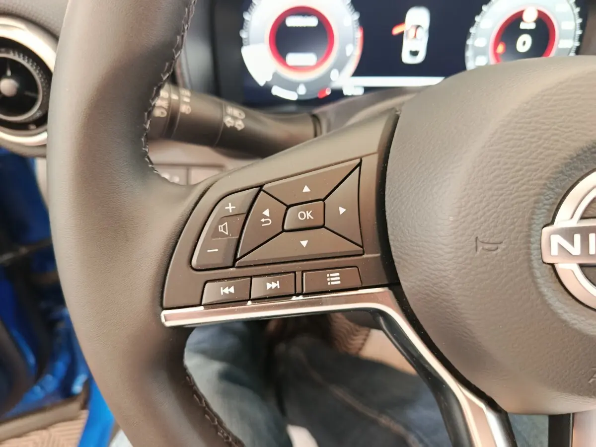 Gros plan sur les commandes gauche du volant cuir du Nissan Juke bleu 2025 avec tableau de bord numérique en arrière-plan.