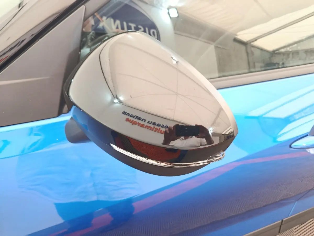 Rétroviseur chromé en gros plan côté gauche d’un Nissan Juke bleu 2025, reflet d’un intérieur d’atelier visible.