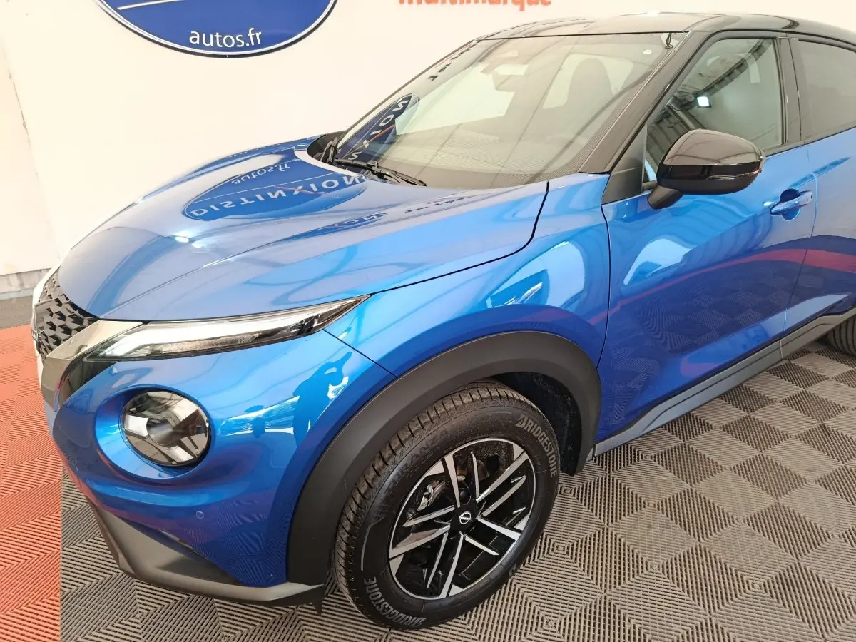 Nissan Juke bleu en 3/4 avant droit, avec phares ronds et jantes noires à motifs argentés.