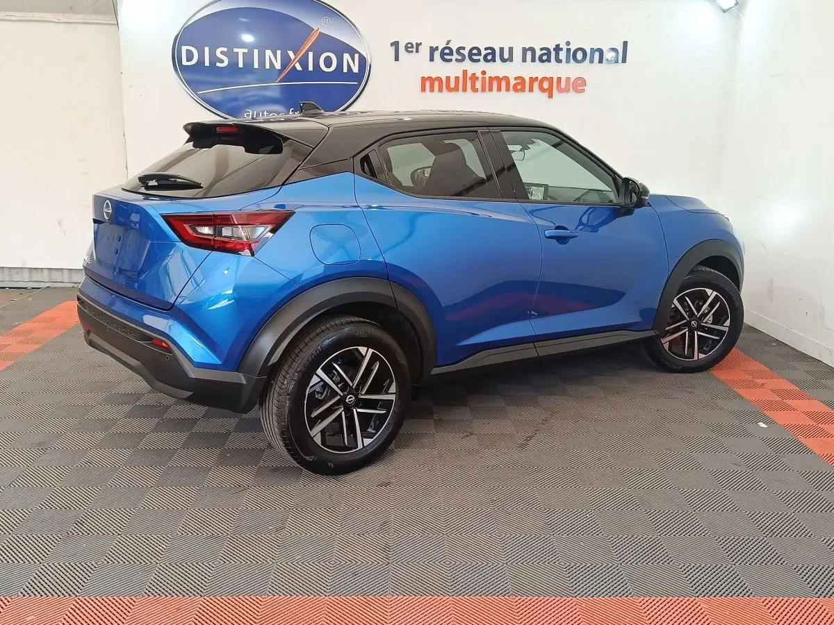 Vue 3/4 arrière droite d'un Nissan Juke bleu 2025 avec toit noir et jantes alliage noires en showroom.