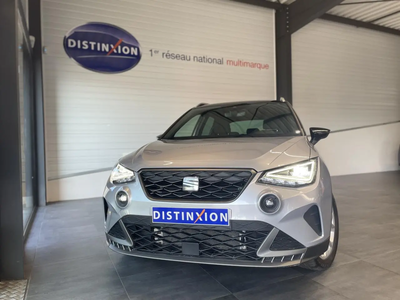Vue avant d'une SEAT Arona 2024 gris clair avec calandre noire et phares allumés en intérieur showroom.