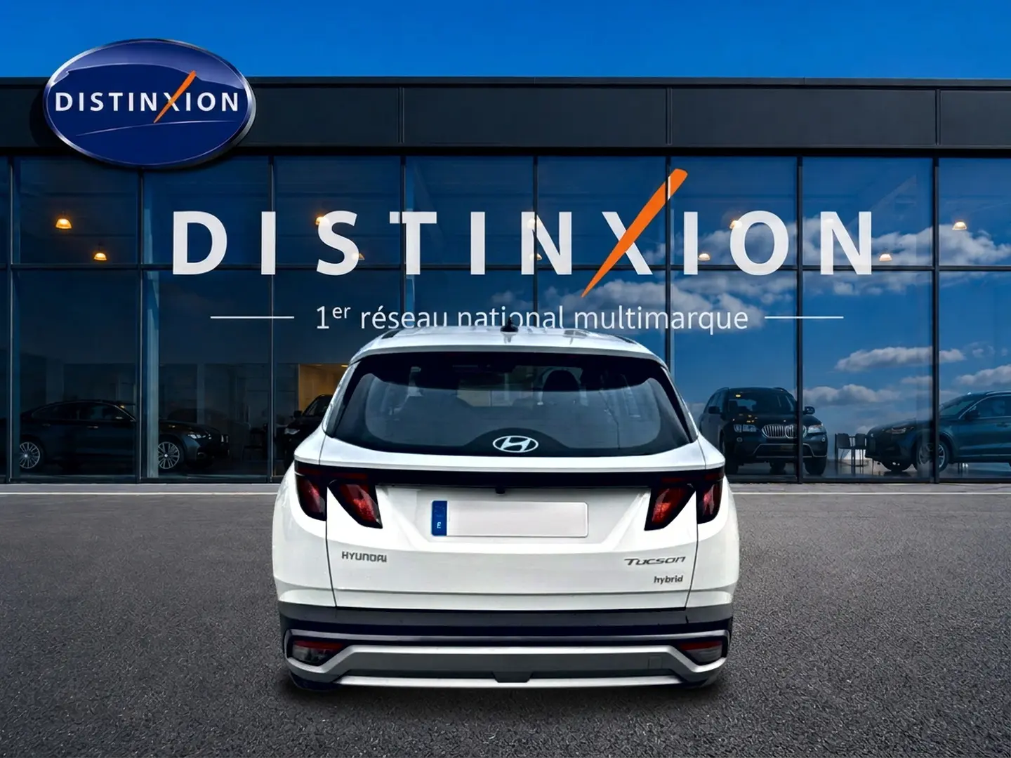 Vue arrière d'un Hyundai Tucson blanc hybride 2026, avec feux sombres et logo Hyundai visible sur un parking devant un bâtiment vitré.