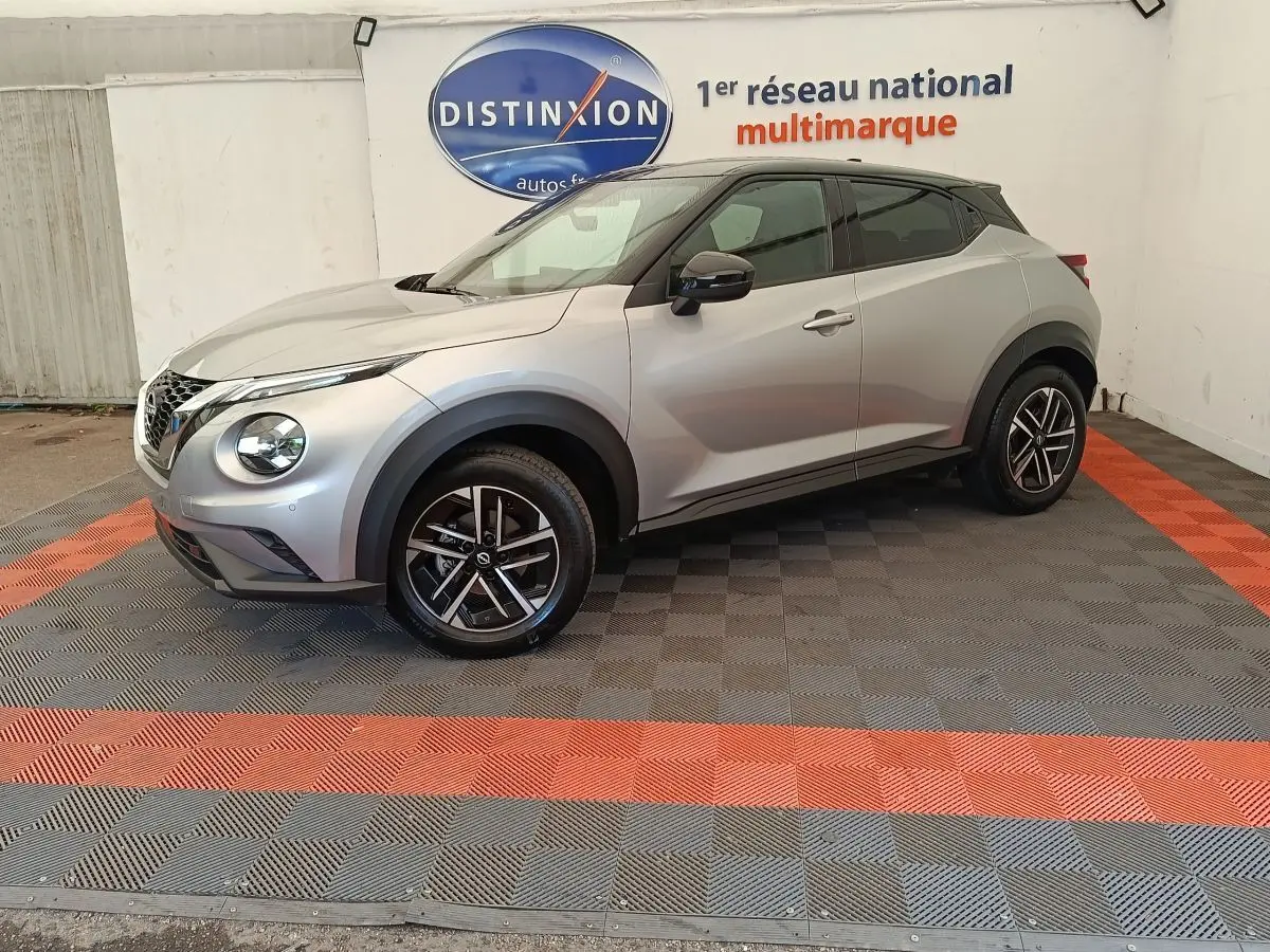 Nissan Juke gris clair vu de profil droit, avec jantes noires et toit noir contrastant, dans un showroom multimarque.