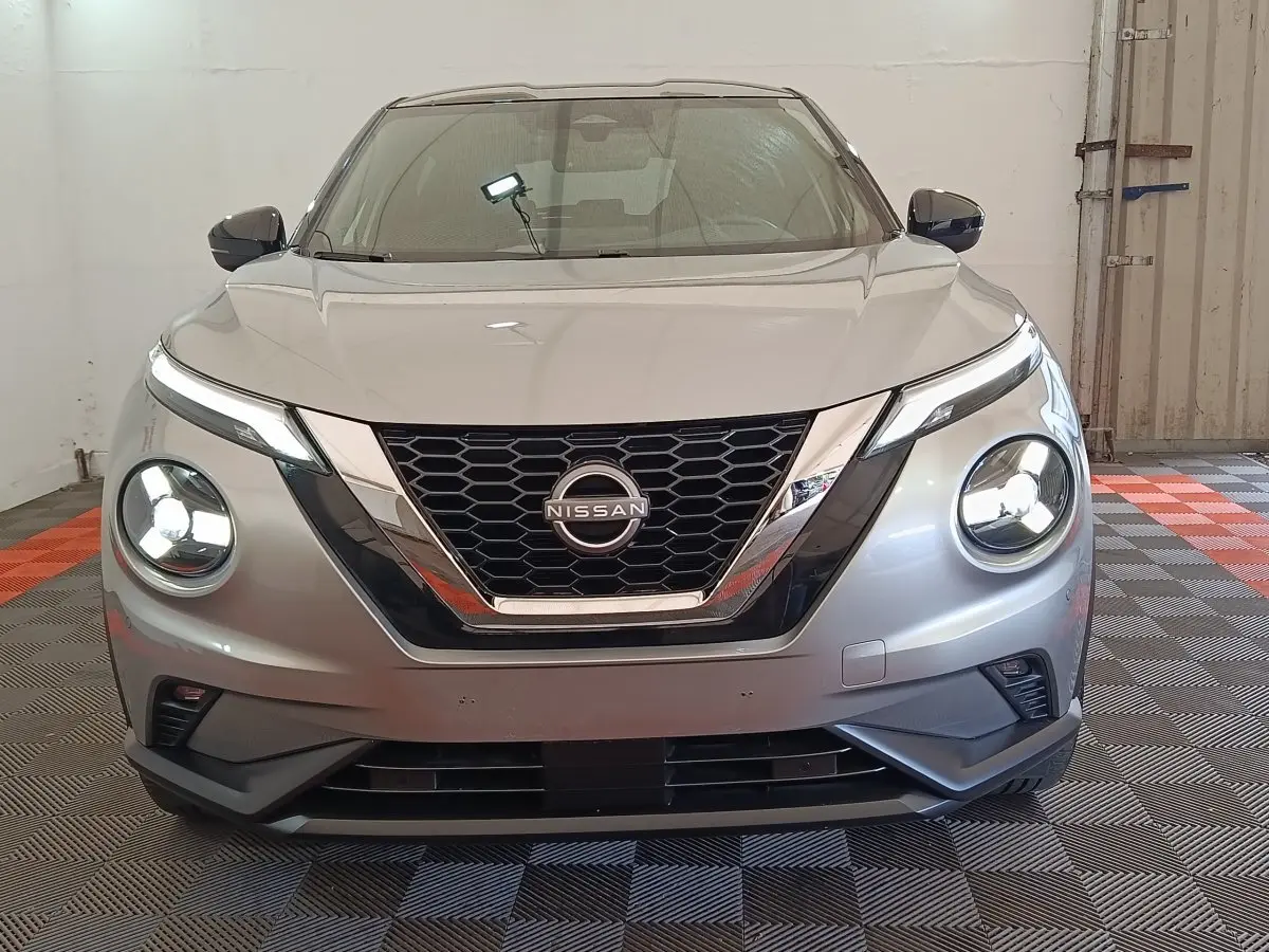 Vue frontale d'un Nissan Juke gris clair 2025 avec phares LED ronds allumés et calandre en nid d'abeille chromée.