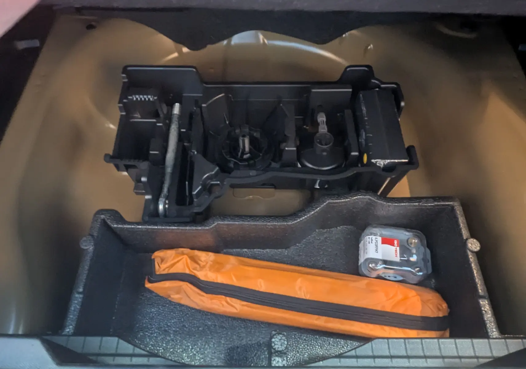 Vue du coffre ouvert d'une Peugeot 2008 noire 2024, montrant le rangement sous plancher avec trousse orange et outils.