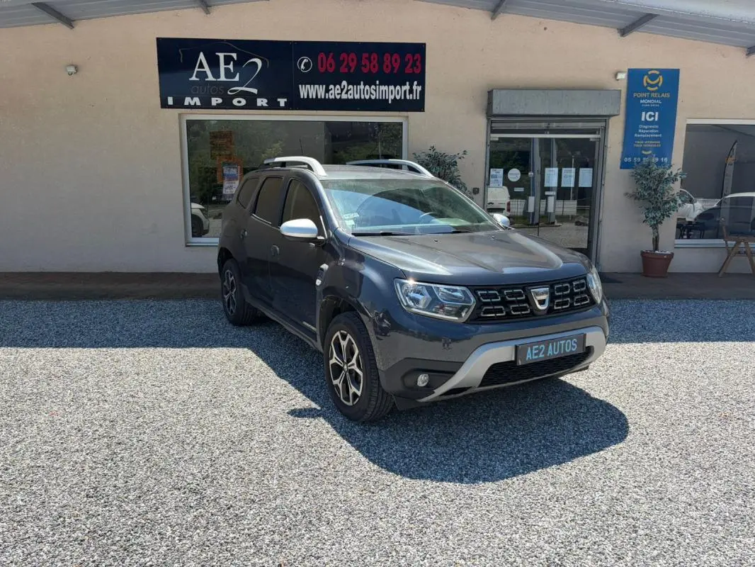 Dacia Duster gris en 3/4 avant droit, avec jantes alliage et barres de toit, stationné devant un garage AE2 Autos.