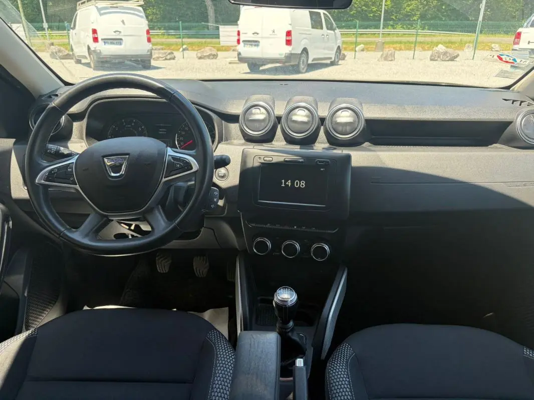 Vue intérieure frontale du tableau de bord du Dacia Duster gris 2021, avec volant, écran tactile et levier de vitesses manuel.
