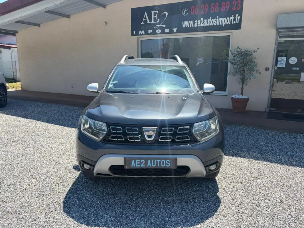 Vue avant d'un Dacia Duster gris 2021 Prestige avec calandre noire et barres de toit visibles, stationné devant un bâtiment.
