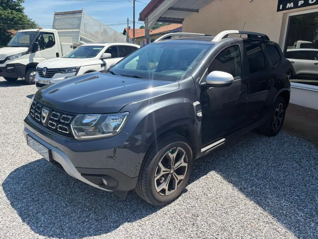 Dacia Duster gris vue 3/4 avant droit, avec jantes alliage et barres de toit visibles, stationné sur gravier.
