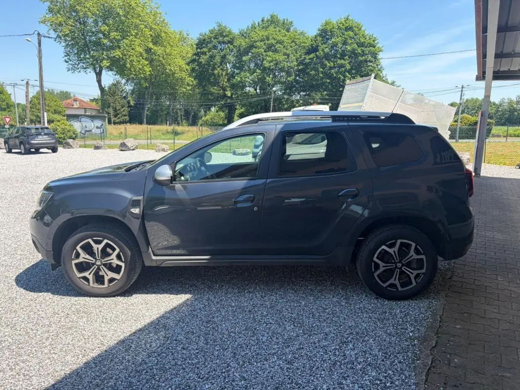 Vue de profil côté gauche d'un Dacia Duster gris 2021 avec jantes alliage et barres de toit noires.