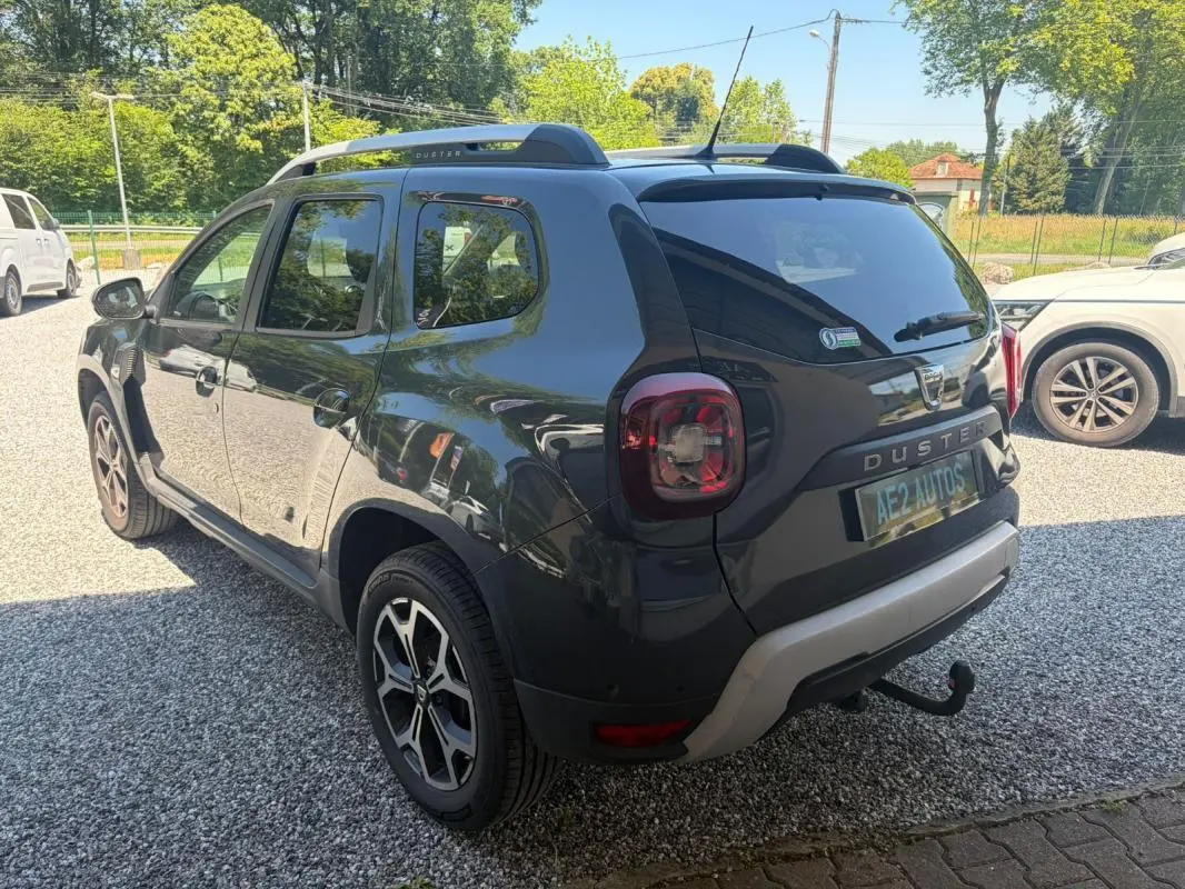 Vue 3/4 arrière droite d'un Dacia Duster gris 2021 avec barres de toit et attelage visible sur un sol gravillonné.