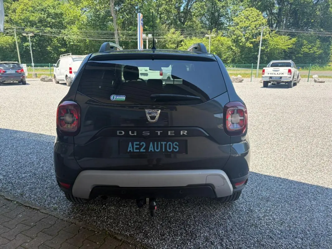 Vue arrière d'un Dacia Duster gris 2021 avec attelage, stationné sur un sol gravillonné en extérieur.