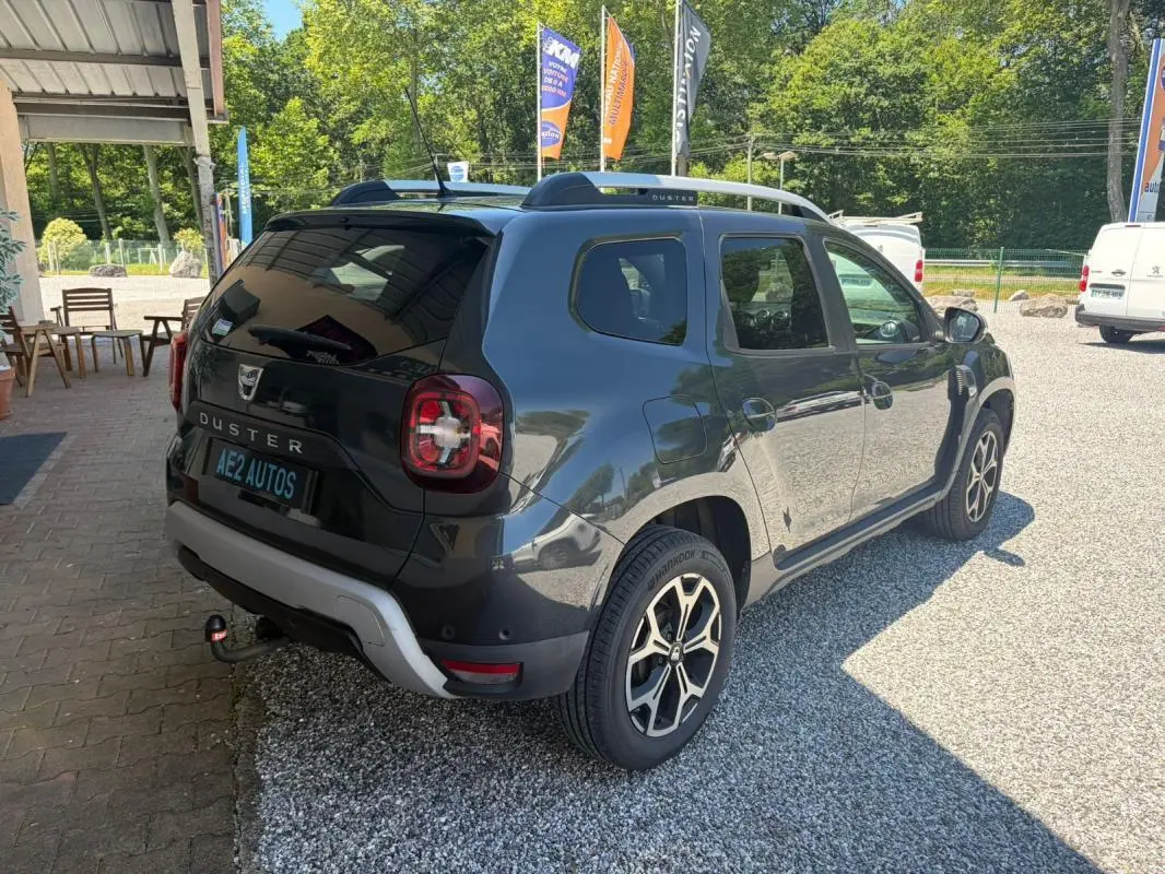 Dacia Duster gris 3/4 arrière droit, avec barres de toit et attelage visible, stationné sur gravier en extérieur.
