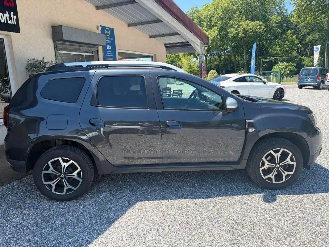 Vue de profil côté gauche d'un Dacia Duster gris 2021 avec jantes alliage et barres de toit argentées.