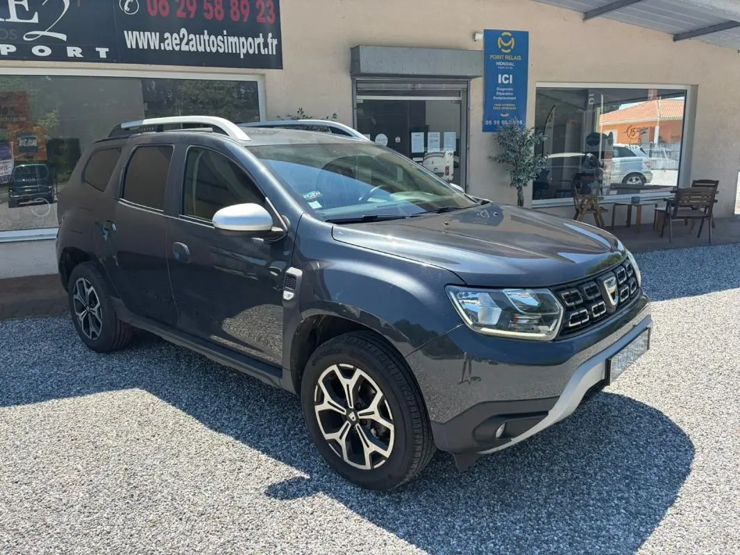 Dacia Duster gris en 3/4 avant droit, avec jantes alliage et barres de toit visibles, stationné devant un bâtiment.
