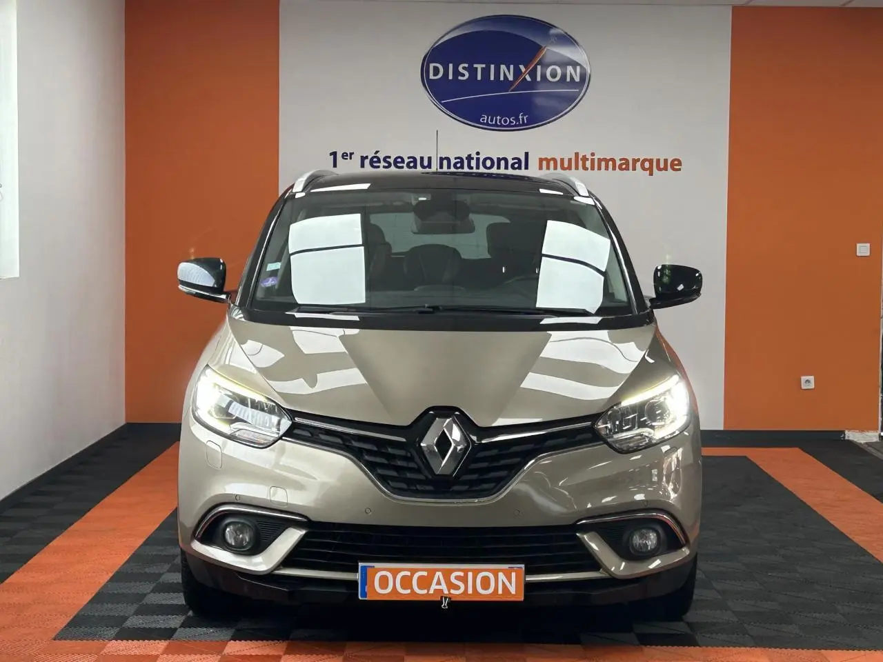 Vue frontale d'un Renault Scénic Grand IV marron clair avec feux allumés dans un showroom orange et noir.