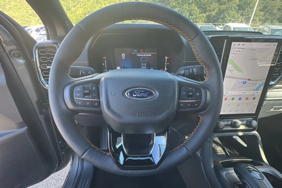 Vue intérieure centrée sur le volant noir avec surpiqûres marron du Ford Ranger 2025 gris carbone, écran tactile et tableau de bord digital visibles.
