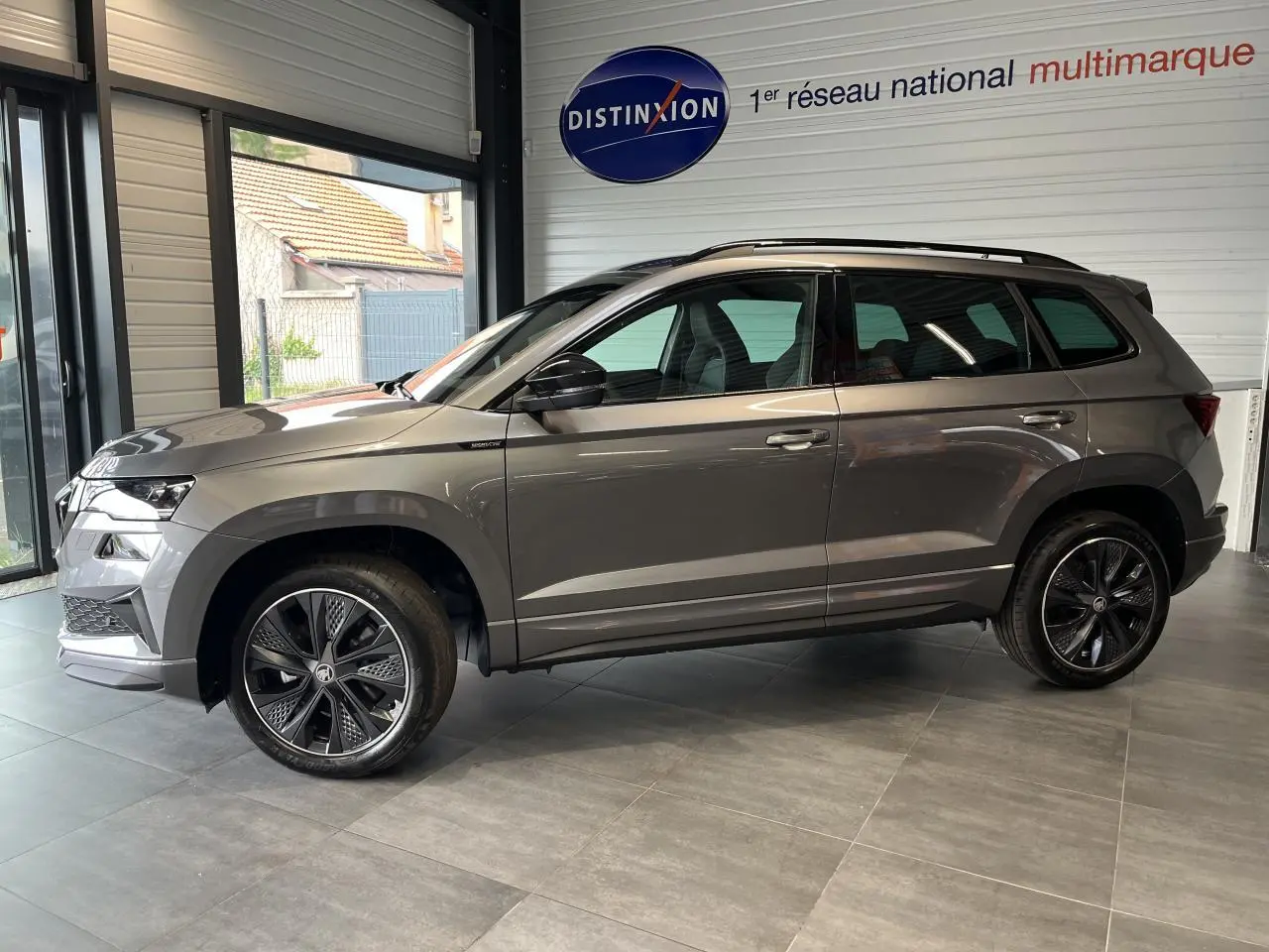SUV SKODA KAROQ gris en profil droit dans un showroom, avec jantes alliage noires et toit noir.