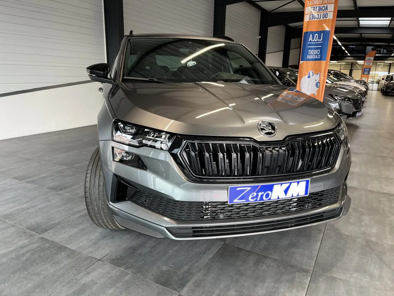 Vue 3/4 avant d'un SUV Skoda Karoq gris métallisé avec calandre noire et phares LED dans un showroom.