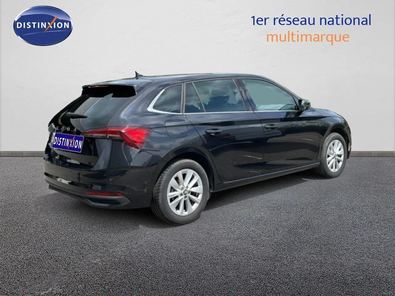 Vue 3/4 arrière droite d'une Skoda Scala noire métal avec jantes argentées, stationnée sur sol gris devant un mur blanc.