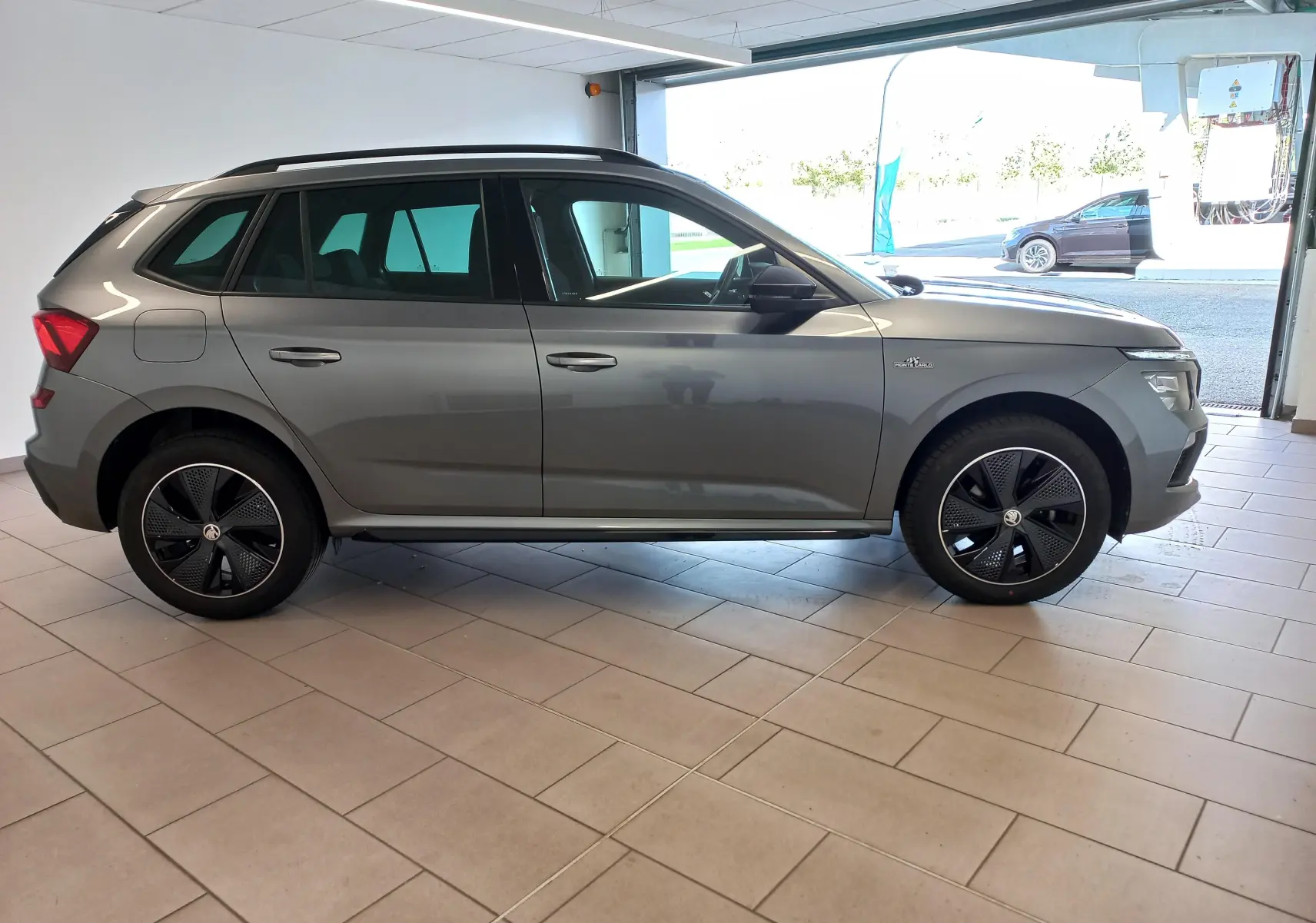 Vue de profil côté gauche d’un Skoda Kamiq Monte Carlo 2024 gris graphite avec jantes noires distinctives en intérieur showroom.
