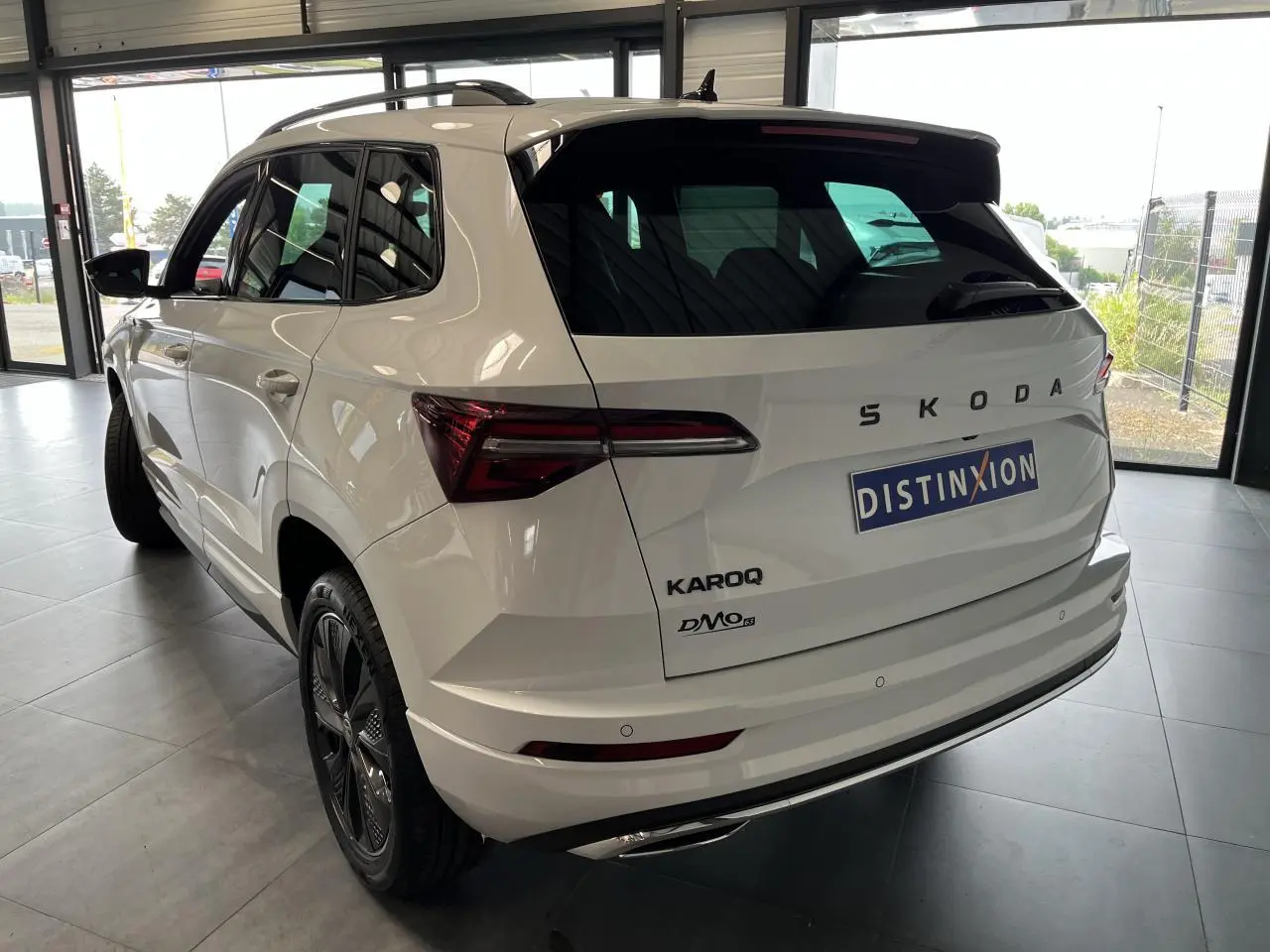 Vue 3/4 arrière droite d'un SUV SKODA KAROQ blanc 2025 avec jantes noires et hayon électrique en intérieur showroom.