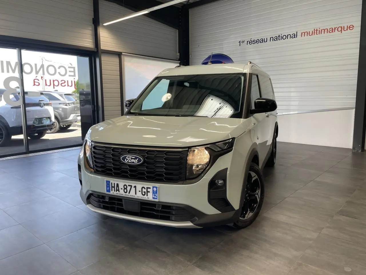 Ford Tourneo Courier gris clair vu en 3/4 avant droit, avec calandre noire nid d'abeille et jantes noires brillantes.