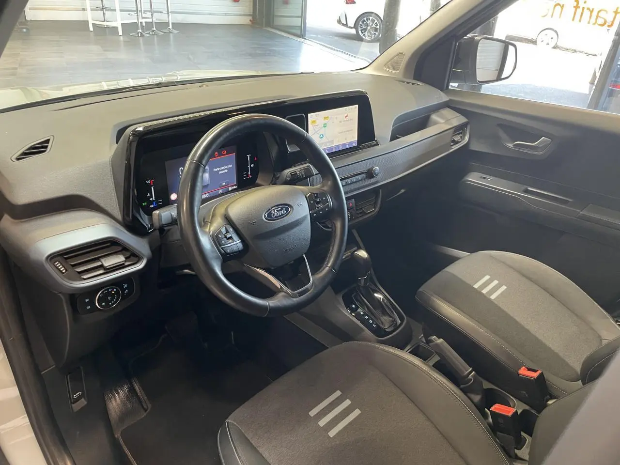 Vue intérieure avant du Ford Tourneo Courier 2024, volant cuir noir, écran tactile 8 pouces et sièges tissu gris foncé.