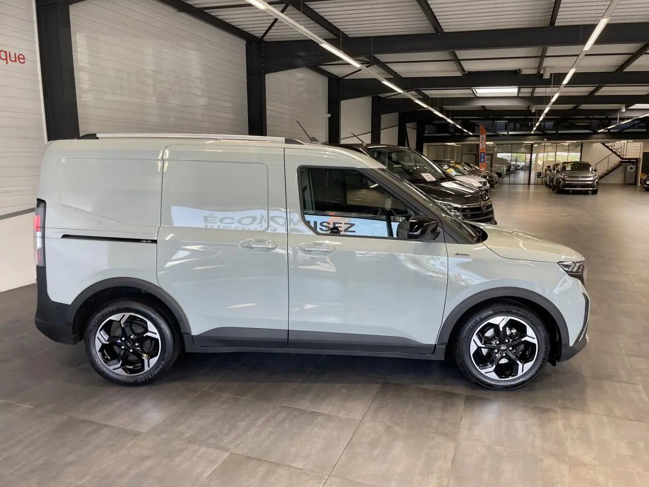 Ford Tourneo Courier gris clair vu de profil côté gauche, avec jantes alliage noires et toit équipé de barres.