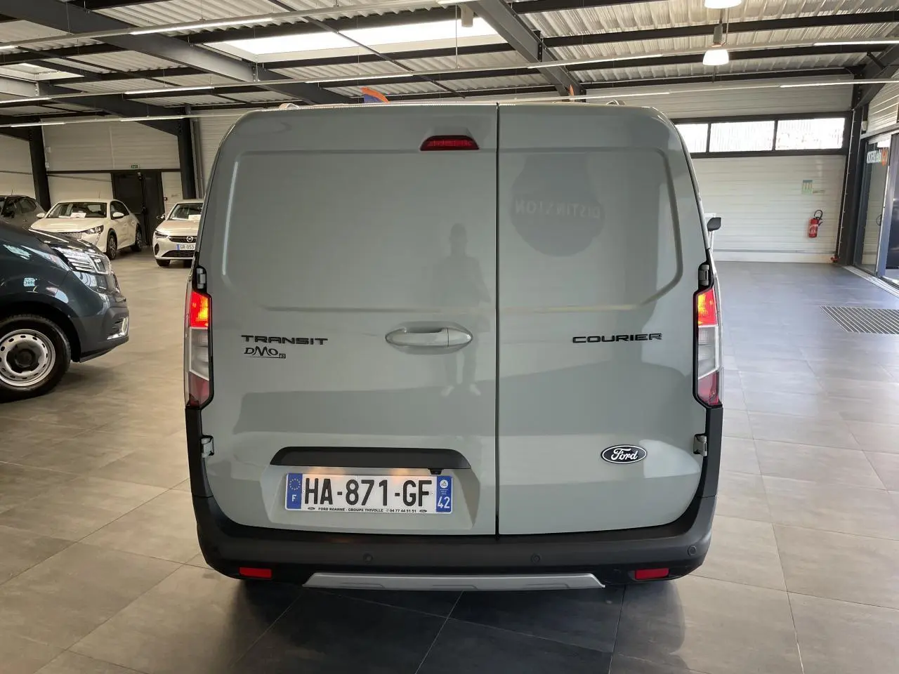 Vue arrière d'un Ford Tourneo Courier gris clair avec portes battantes et feux arrière allumés en intérieur showroom.