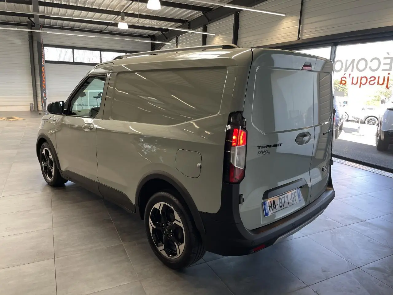 Vue 3/4 arrière droite du Ford Tourneo Courier gris clair avec jantes aluminium et feux arrière allumés en intérieur showroom.