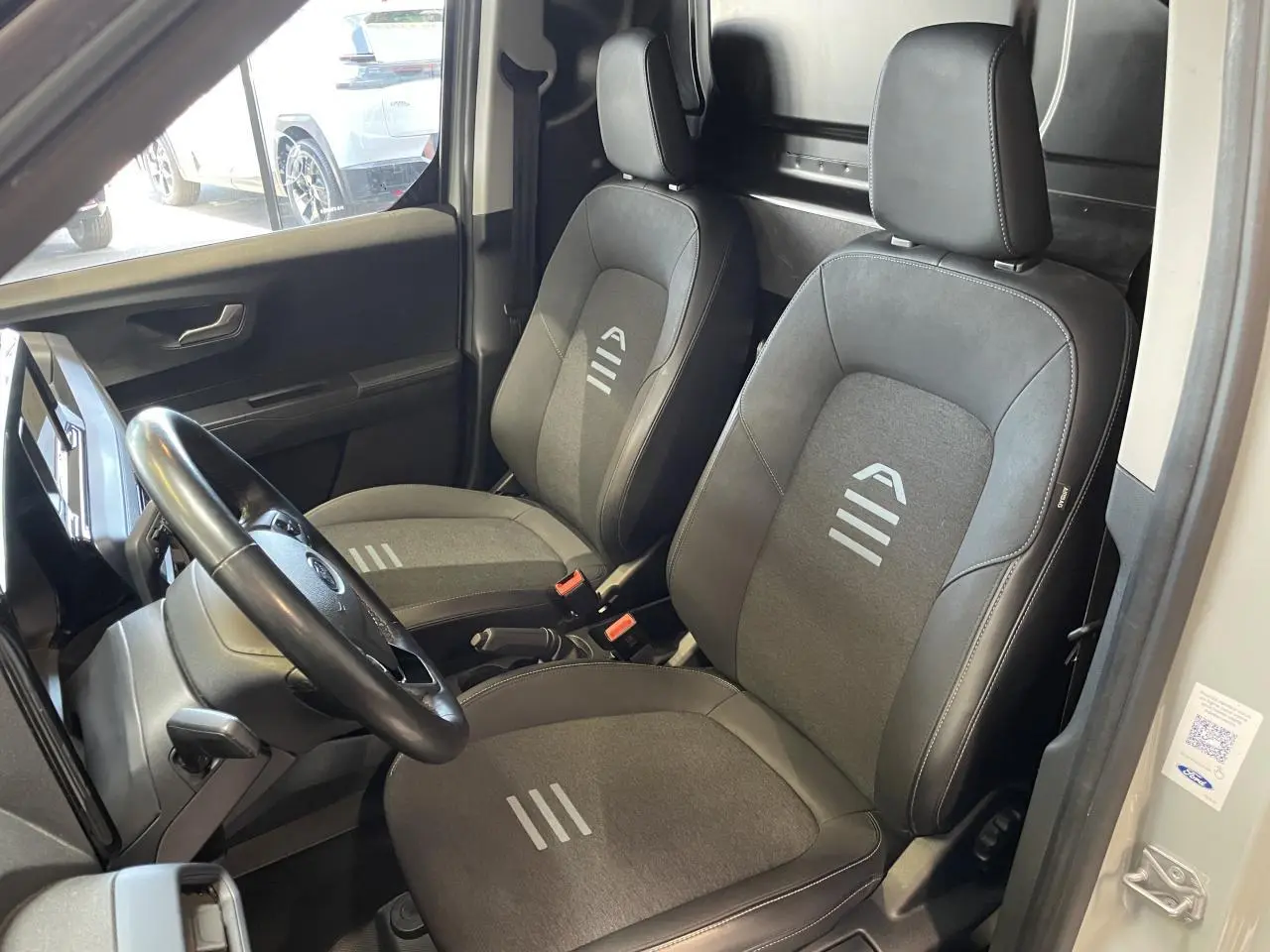 Vue intérieure côté conducteur du Ford Tourneo Courier gris clair 2024, sièges tissu avec logo et volant cuir synthétique.