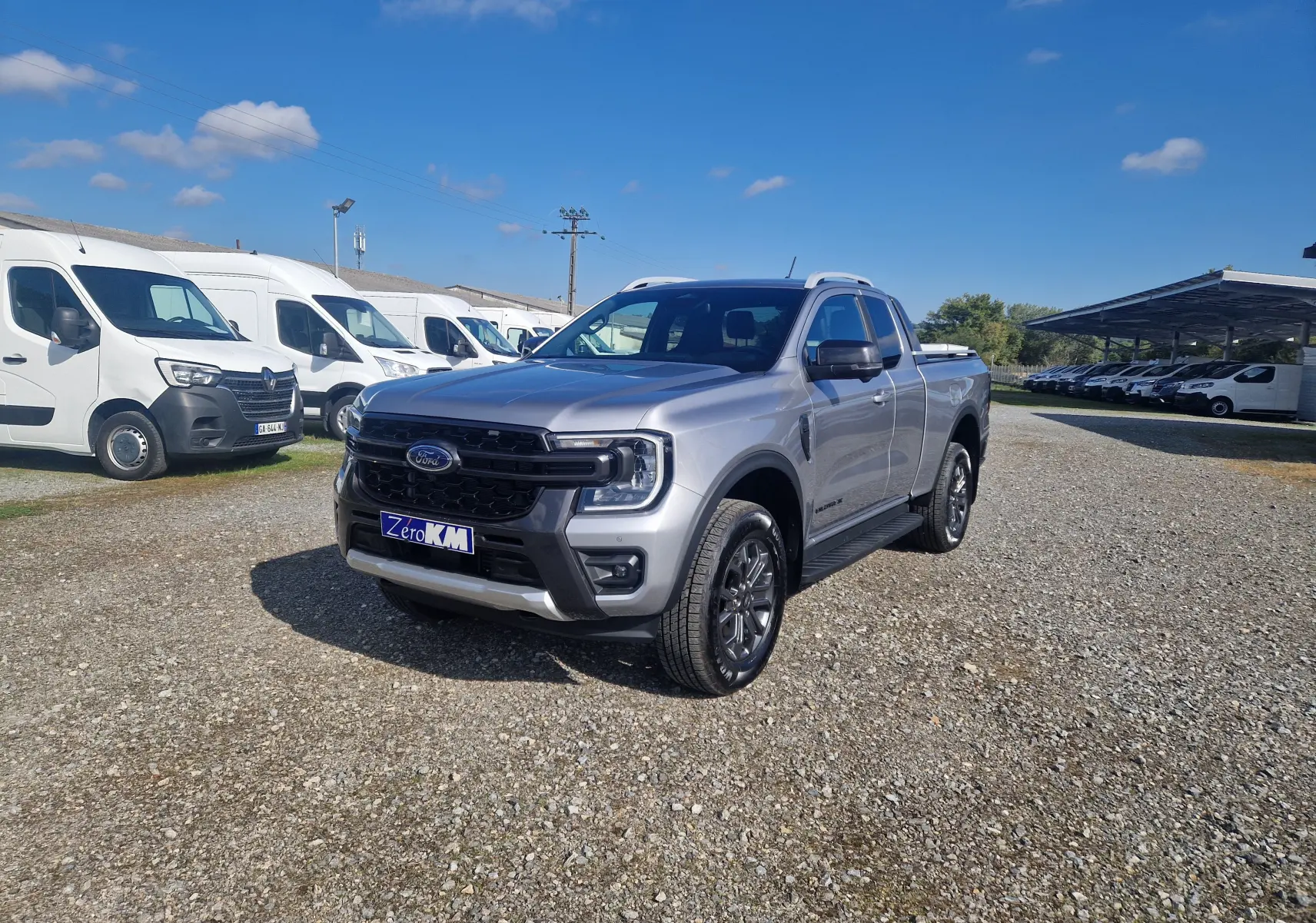 Ford Ranger Super Cabine gris Iconic vu en 3/4 avant droit sur un parking extérieur sous un ciel bleu.