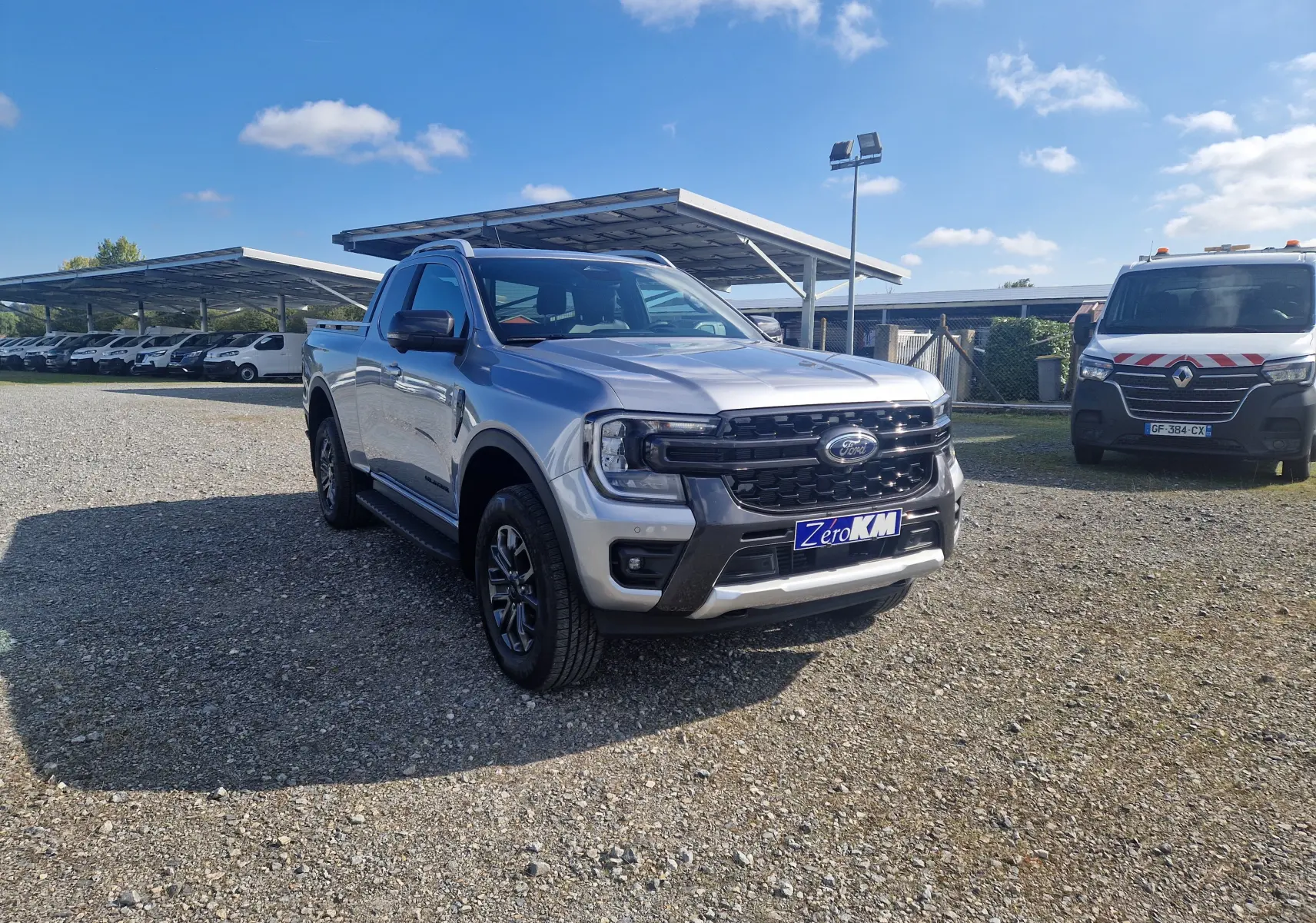Ford Ranger Super Cabine gris iconic en 3/4 avant droit sur un terrain gravillonné avec plusieurs utilitaires blancs en arrière-plan.