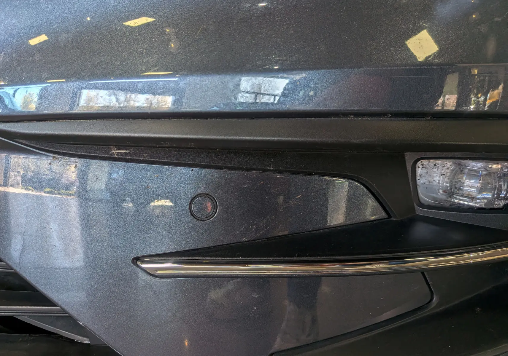 Gros plan sur le pare-chocs avant gris titanium de la Renault Mégane IV Berline avec capteur de stationnement et antibrouillard.